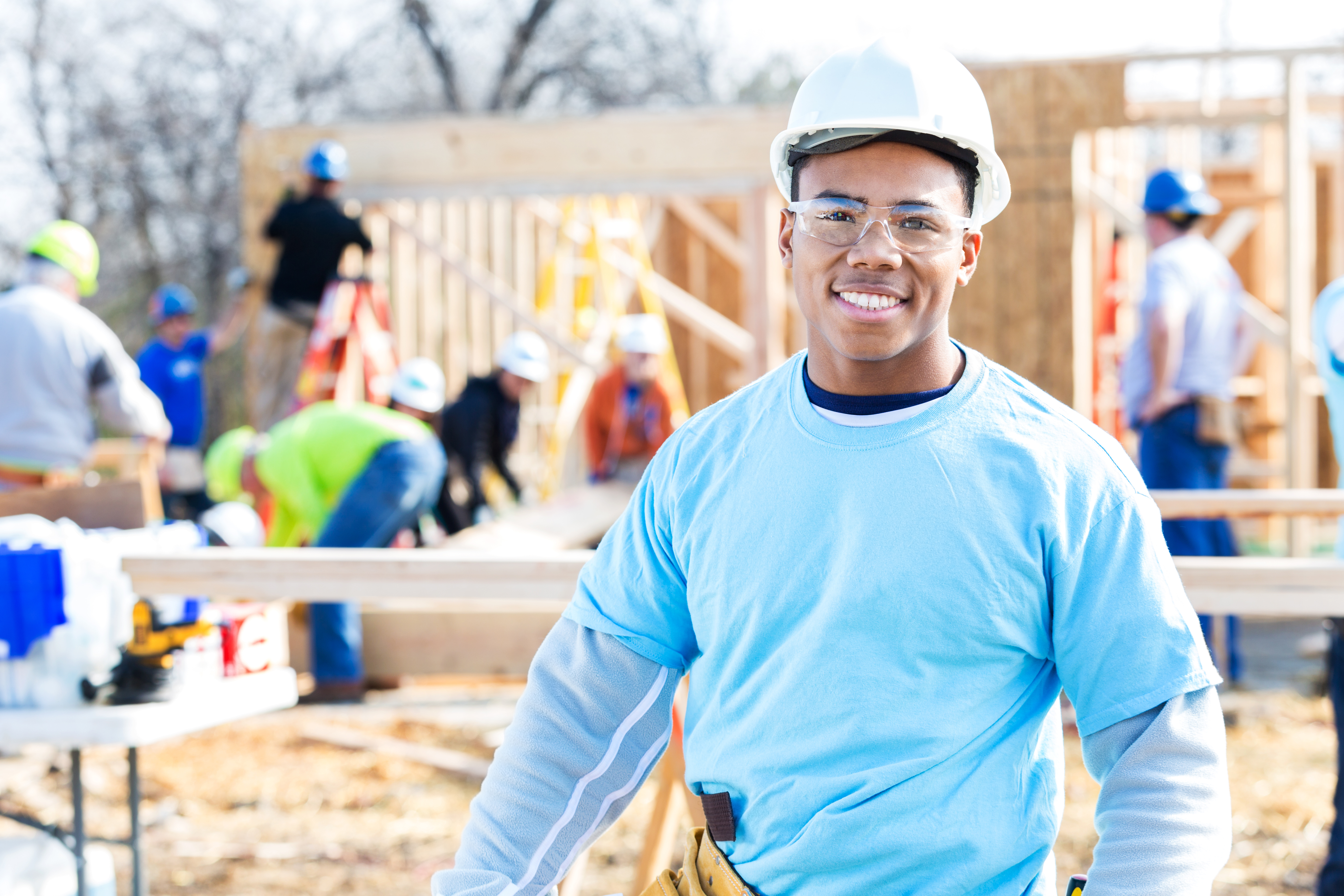 Handsome African American construction foreman or building contractor stands in front of home that is being built for charity. Volunteers are working on the home in the background. He hsi wearing a hard hat, safety glasses and volunteer t-shirt.